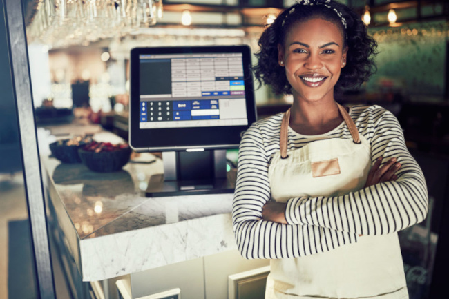 Cheerful woman in a striped shirt and apron stands in front of a restaurant's point of sale system, representing the integration of operational processes in hospitality.